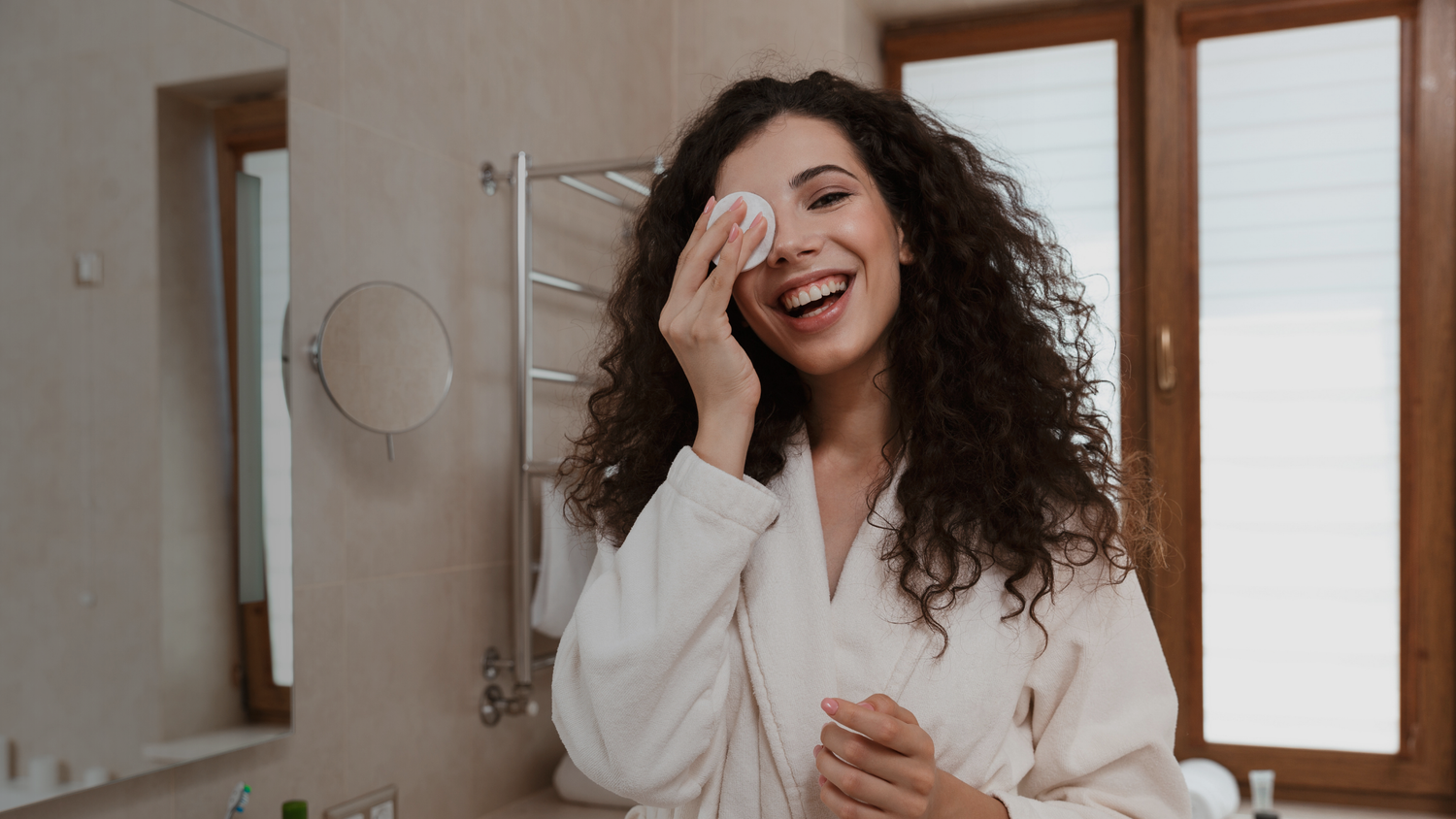 Woman in a bathrobe applying cream to her face in a bathroom.