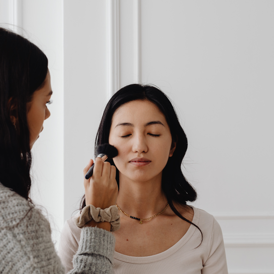 Two women with one applying makeup to the other against a white wall.