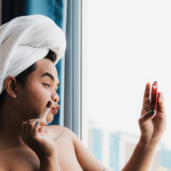 Person applying makeup in front of a window with a towel on their head